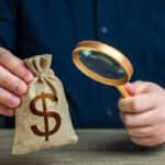 A man examines a dollar money bag through a magnifying glass. Investigating capital origins. Anti money laundering, tax evasion. Study terms and conditions on deposit or loan. Find investments