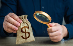A man examines a dollar money bag through a magnifying glass. Investigating capital origins. Anti money laundering, tax evasion. Study terms and conditions on deposit or loan. Find investments